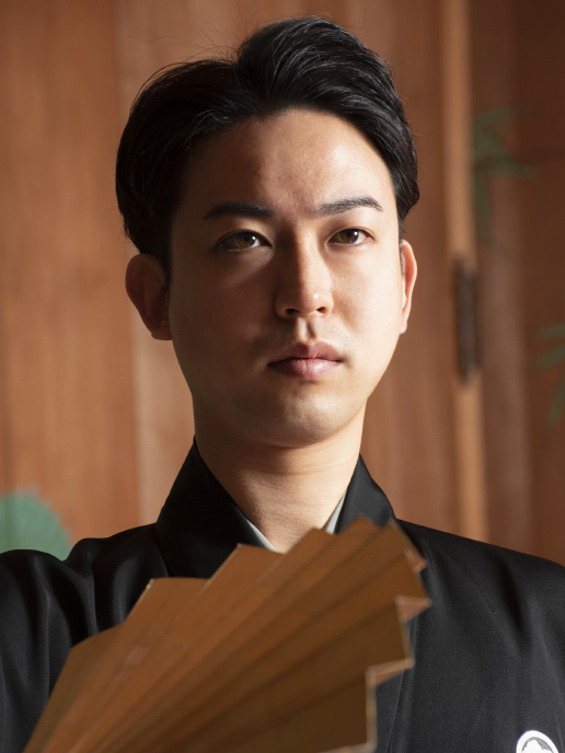 Kazuma Tatsumi in crested black kimono, holding a fan