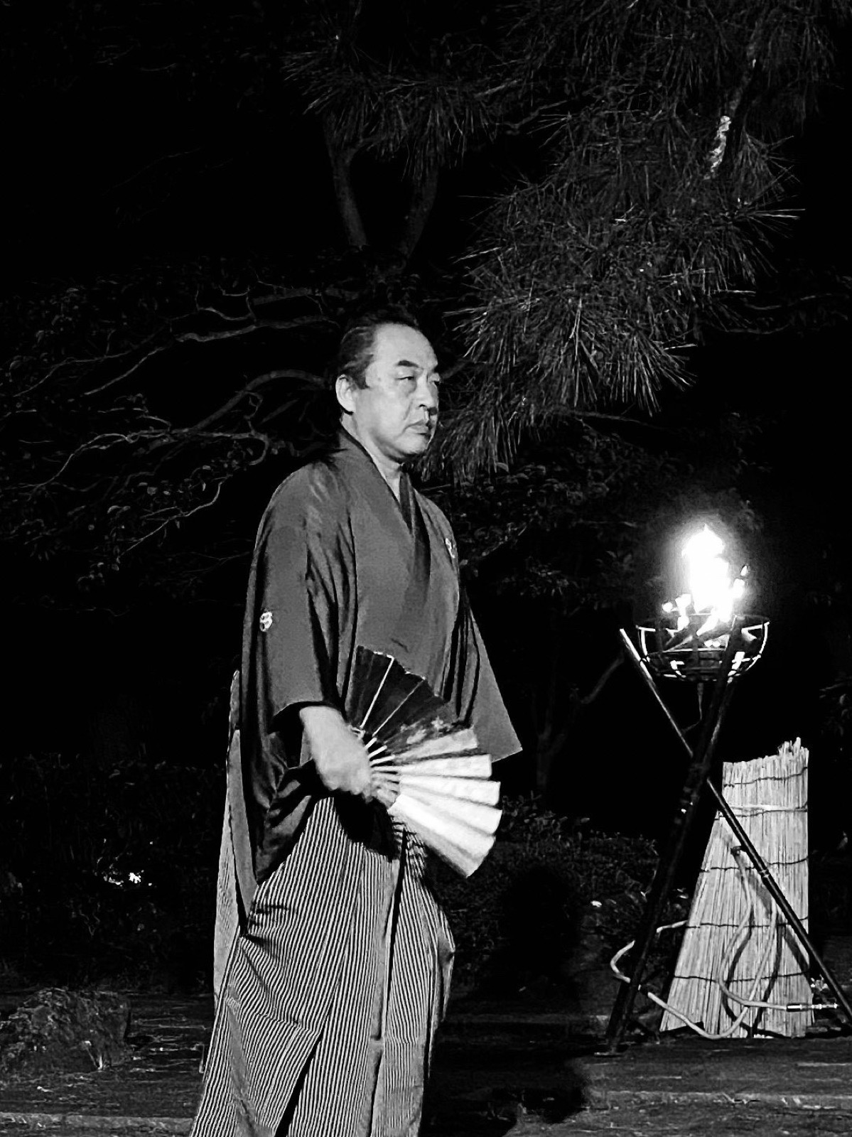 Manjirō Tatsumi in formal kimono, holding a fan beside a torch